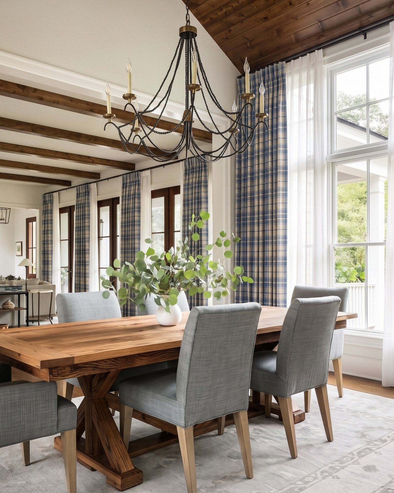 A dining room with blue and cream plaid curtains layered over white sheers, a rustic wooden table, gray upholstered chairs and an iron chandelier