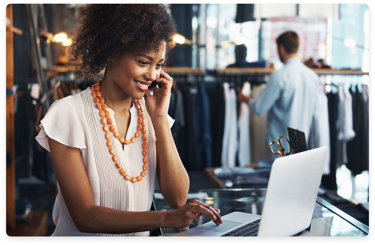 Smiling businesswoman using a laptop and speaking on the phone in a fashion boutique, representing a client-focused approach to financial health and proactive management in a retail setting.