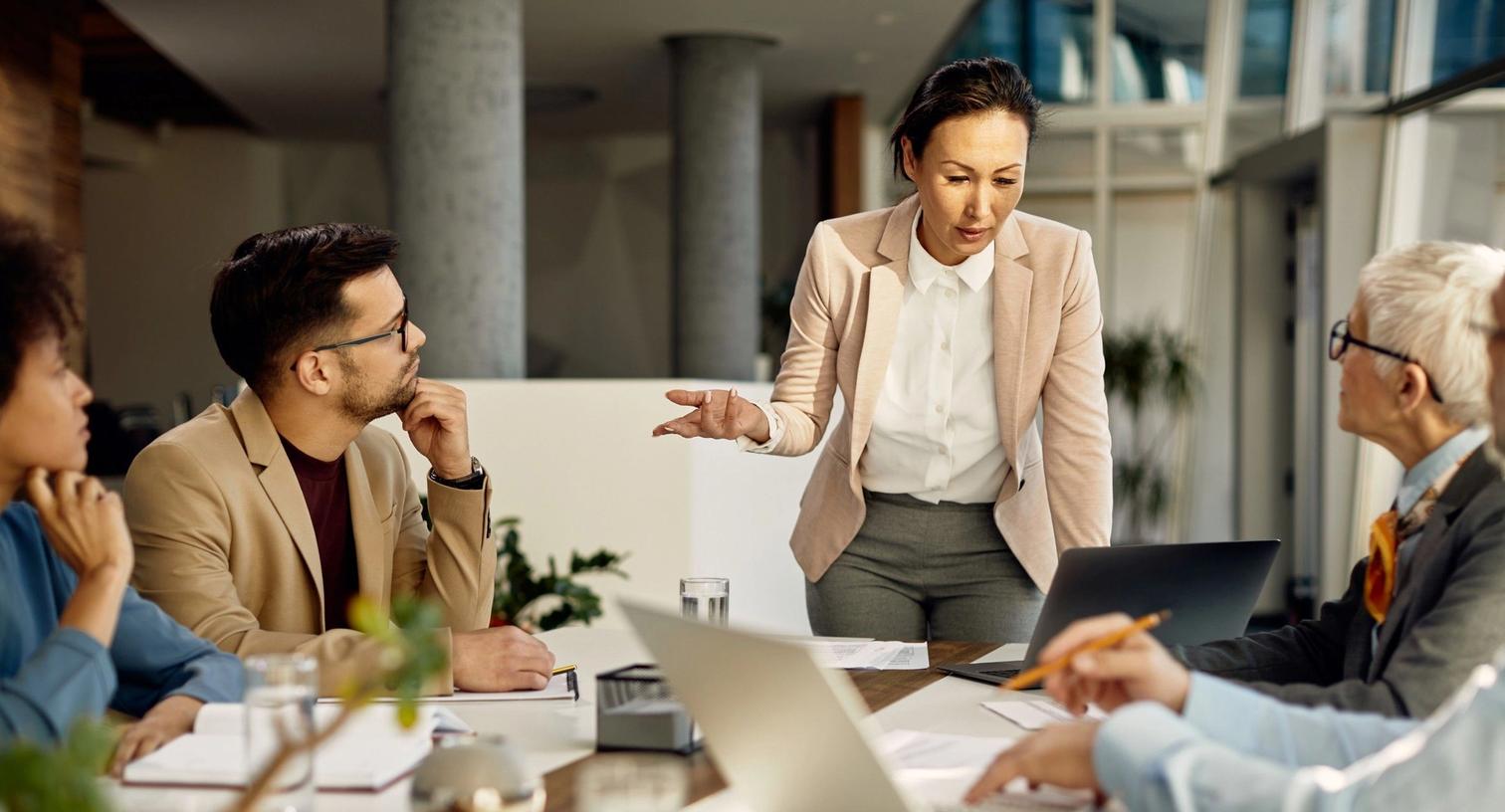 A woman stands presenting at the end of a conference table to two attentive colleagues, in a modern, well-lit office, representing a professional business meeting.