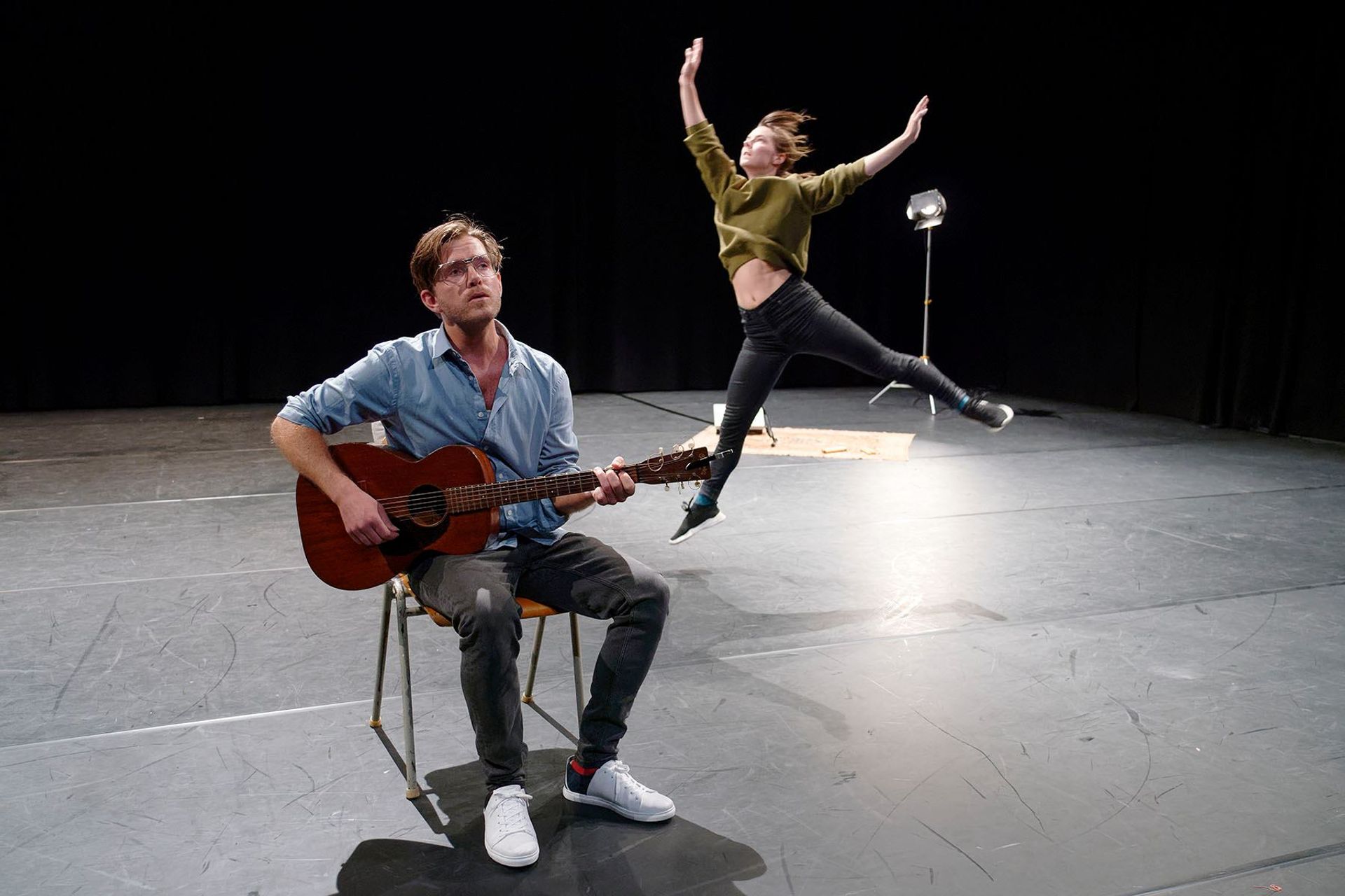 Lasse is seated on a chair with a guitar on his lap, singing and playing. Ingrid is in the background, jumping in the midst of a dance.