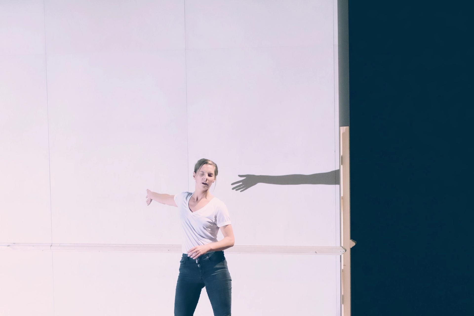 Ingrid is standing on stage in front of a wall that serves as a backdrop. Her eyes are closed and her arms is stretched out, casting a shadow on the wall behind her. 
