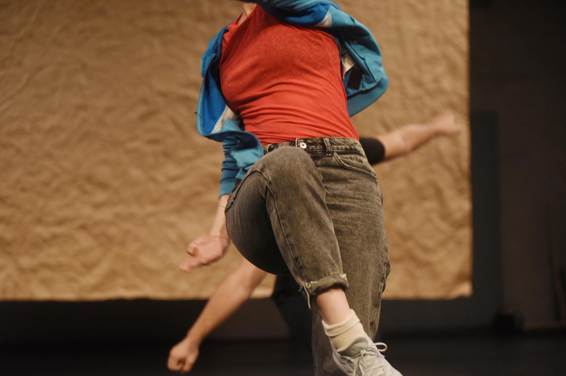 Ingrid is dancing, is wearing a red shirt, jeans and a blue adidas jacket. Her torso is visible against a brown wringkled paper backdrop. 