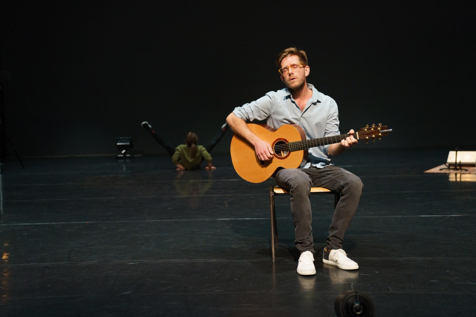 Lasse is in the foreground sitting on a chair, playing a song on the guitar. His eyes are closed and he is singing. In the backgroudn, Ingrid is in the middle of a dance. 