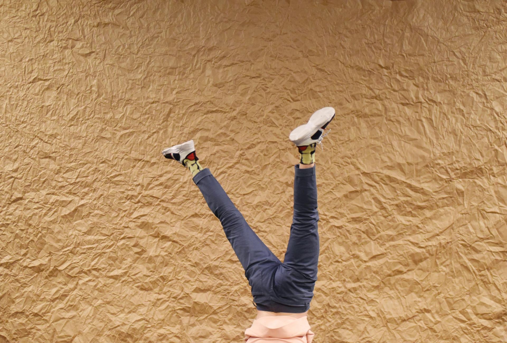 Pablo is doing a handstand, only his plue pants and sneakers visible in the frame, against a brown wrinkled paper backdrop. 