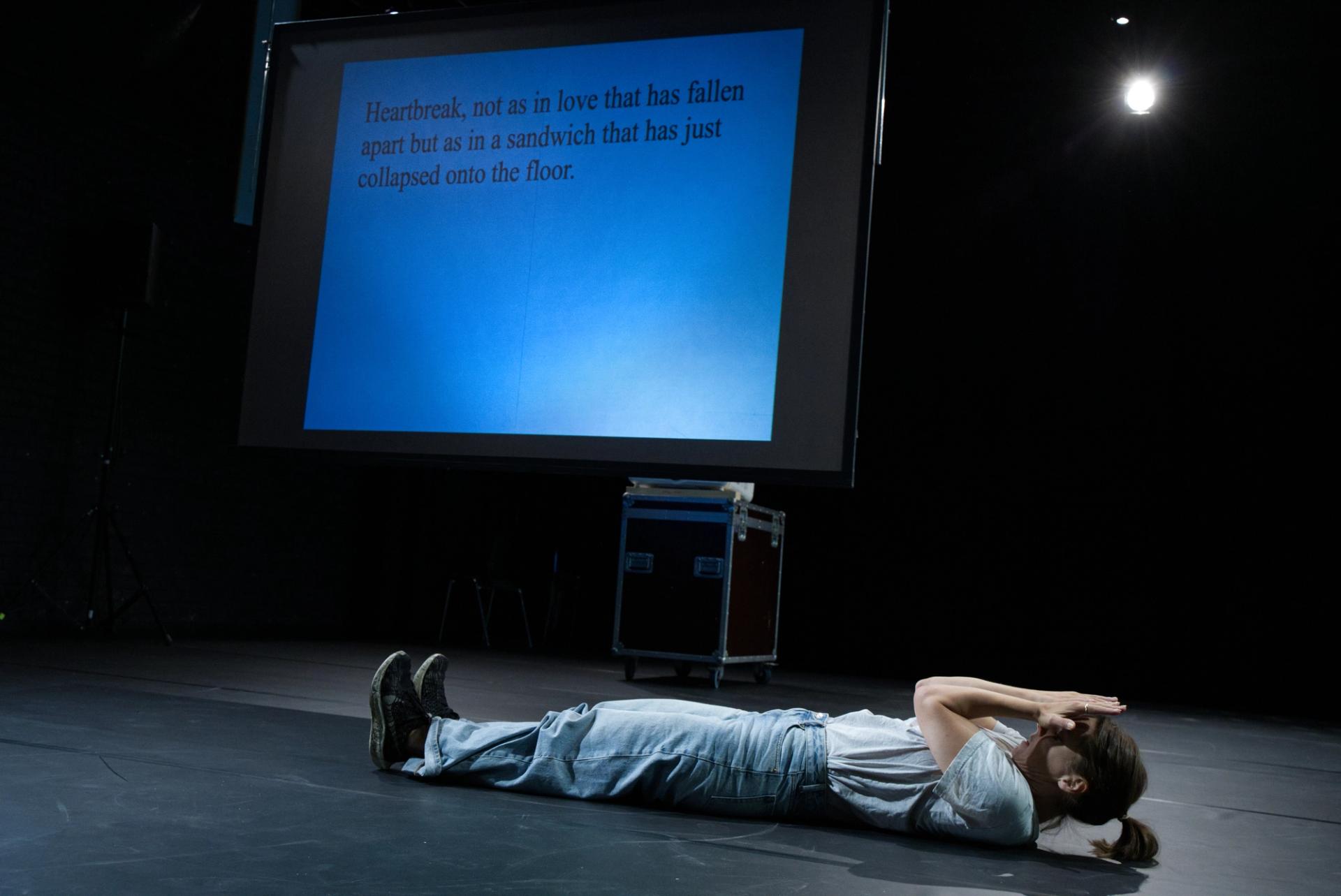 A caption in the font times new roman is projected brightly on the back wall of a small room, describing a dance. Next to it, Ingrid is in mid-motion dancing the dance described.