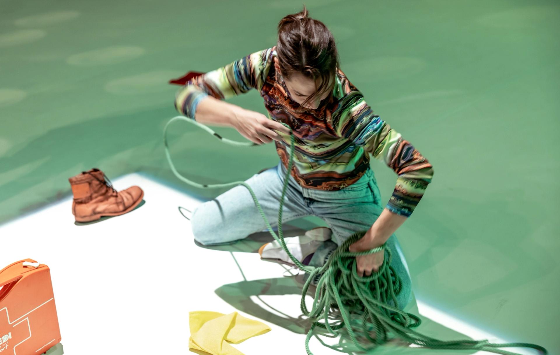 Ingrid is sitting on the floor in pale jeans and a colourful shirt. Around her are several mundane objects and she is managing a green rope. 