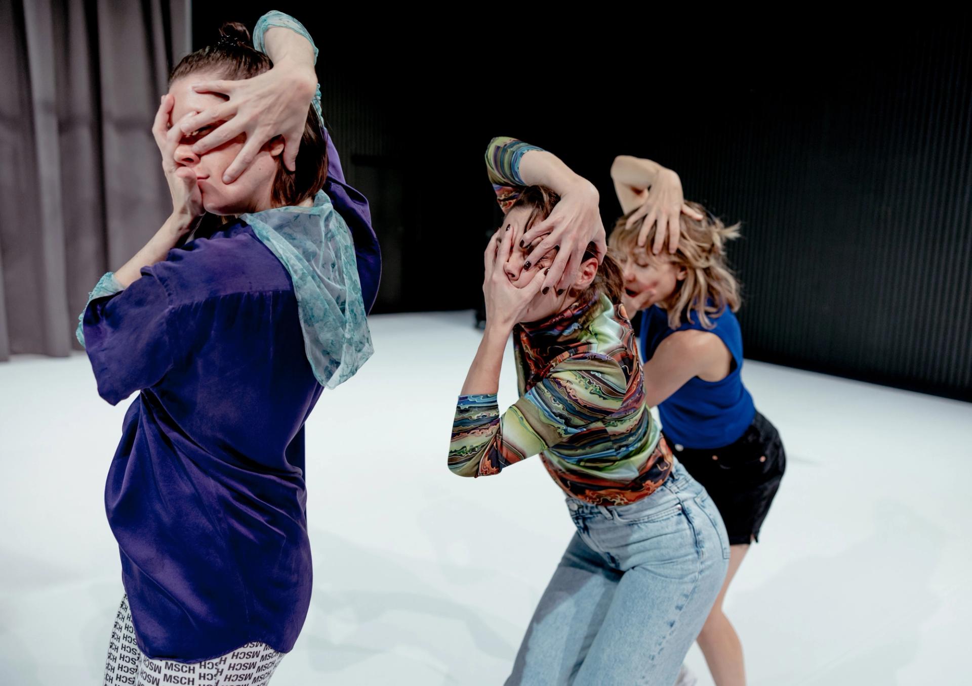Ingrid, Nicola and Ida in a line on a white an grey stage. Their hands are twisting, covering their faces. 