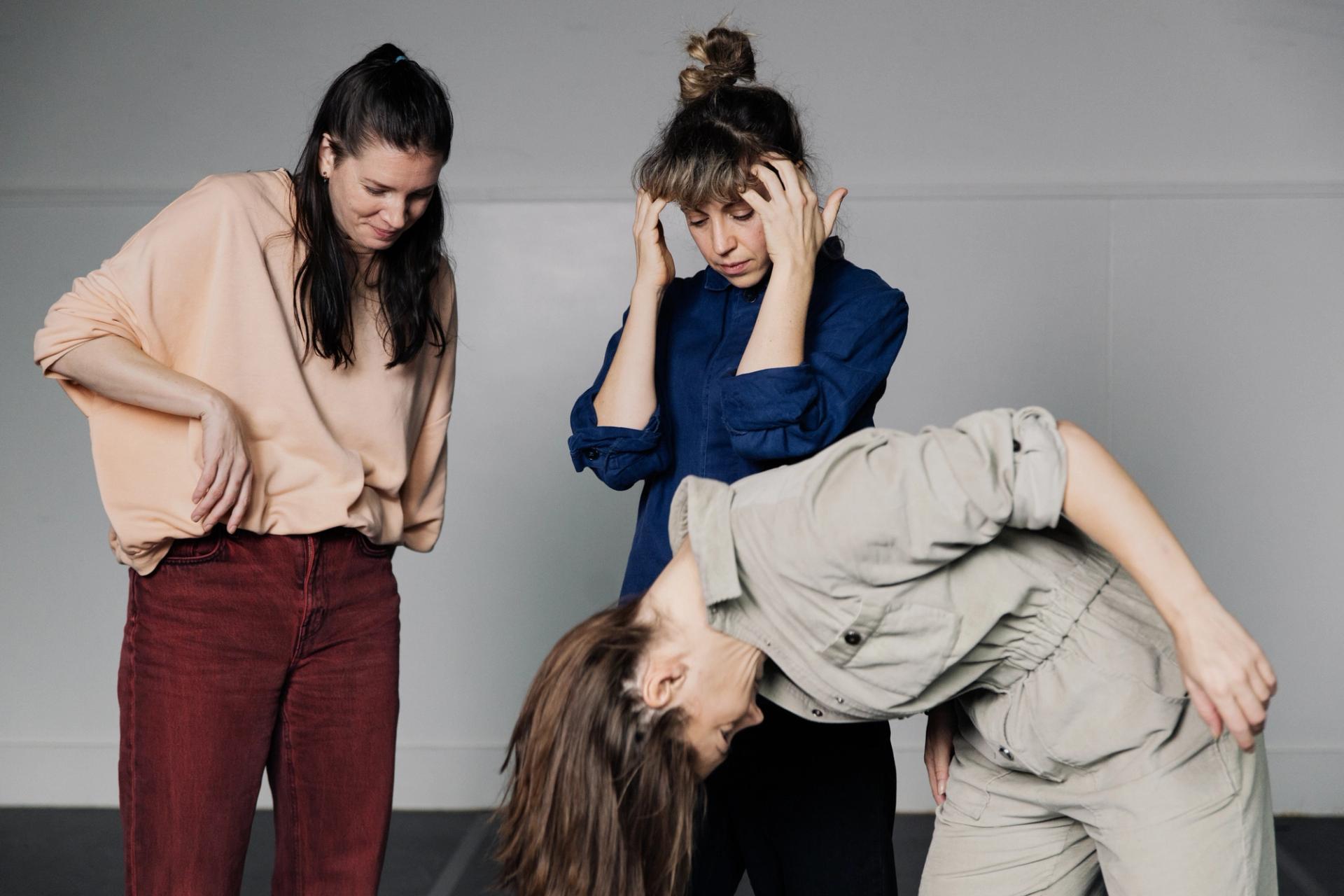 Ida, Nicola and Ingrid in a studio against a grey back wall. They are in the middle of a readjustment movements, Ingrid bending over to shake her hair, Ida correcting her short and Nicola with her hands gesturing towards her head as if thinking. The atmosphere is light. 