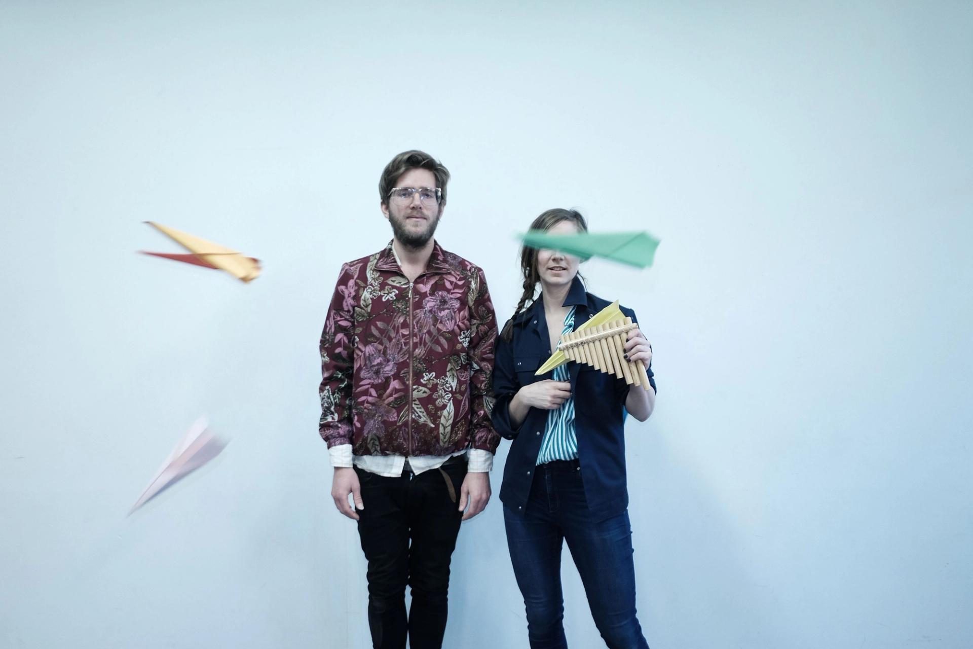 Lasse and Ingrid are standing smiling against a light blue backdrop. Ingrid is holding a panflutes and around them are flying paper planes, one of which happens to partially cover Ingrid's face. 