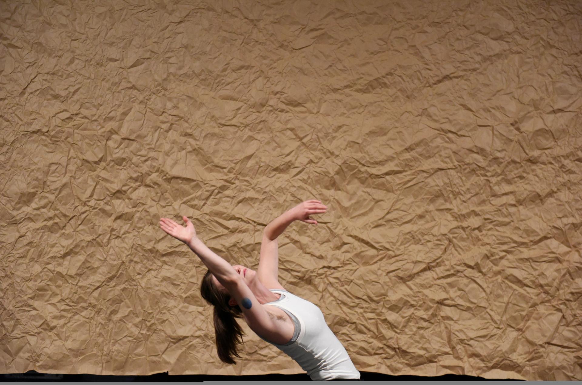 Chloe is leaning backwards, her hand covering her face in the middle of a dance. Only her upper body is visible, against a wrinkled  brown paper backdrop. 