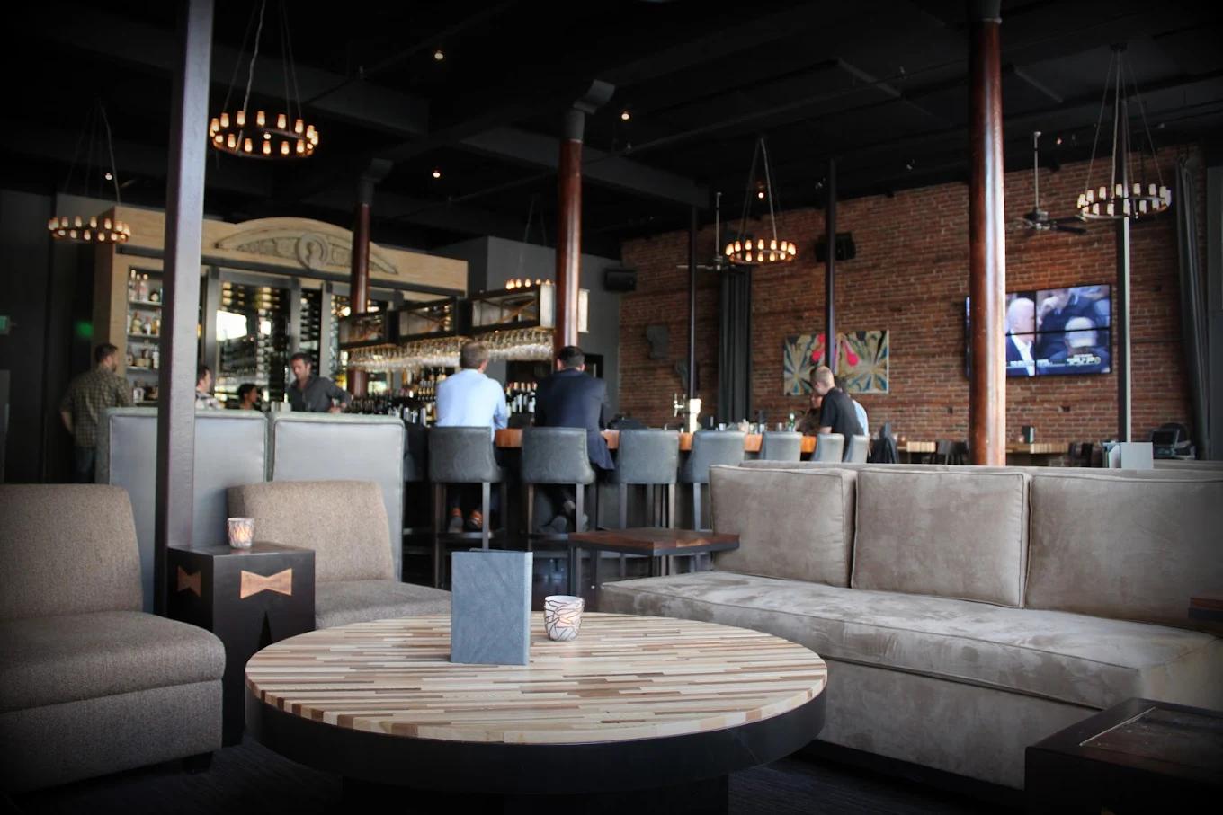Couches sit in the foreground as two well-dressed men eat food at a bar in the background