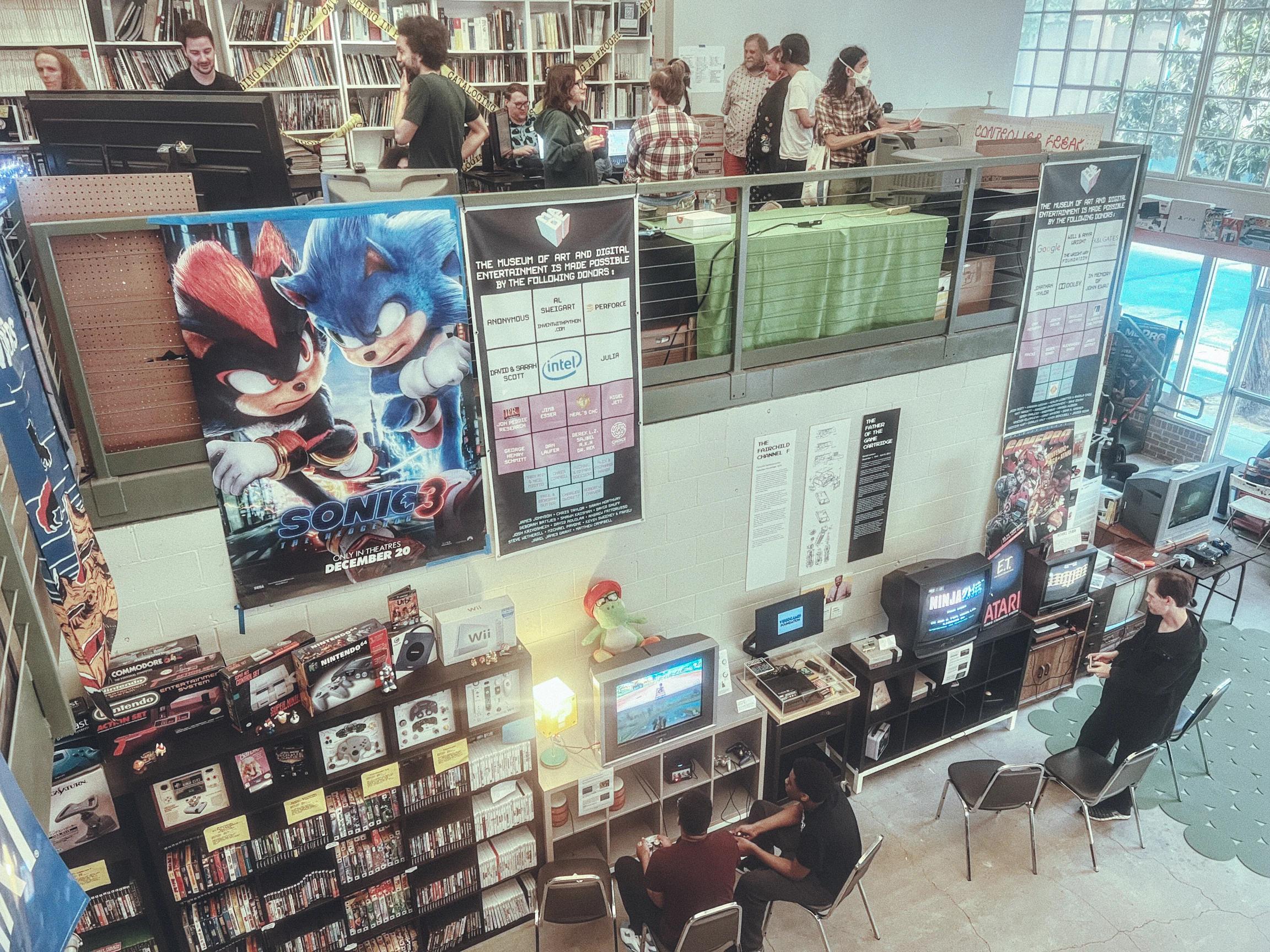 A videogame museum with two floors has visitors playing below and showcasers showing games on the top floor