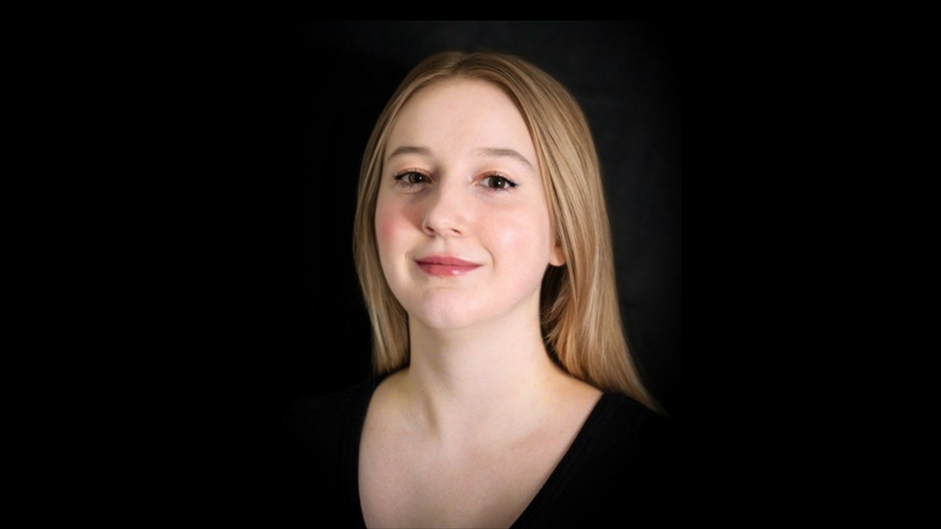 A picture of a young blonde woman looking at the camera, she is smiling. The background is black.