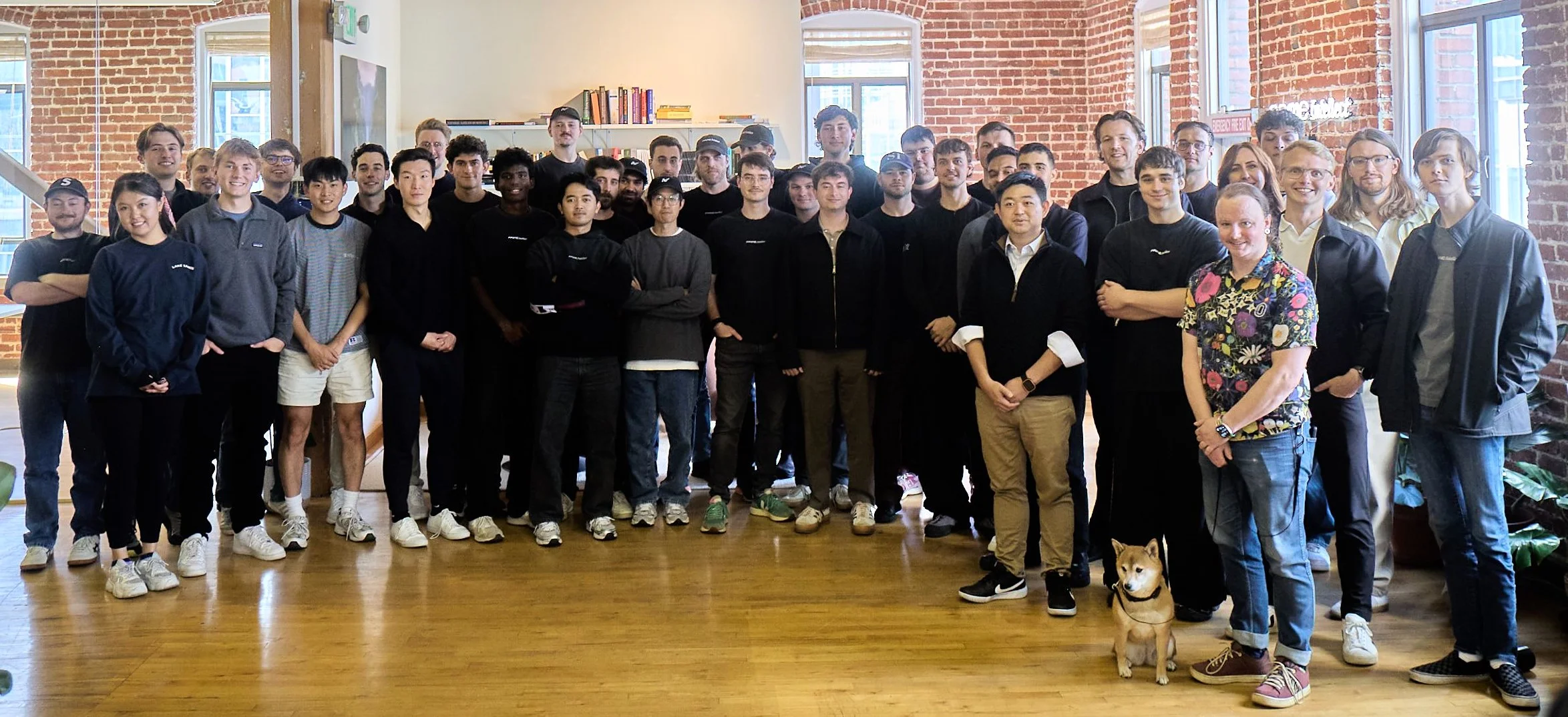 A group photo of about 40 people standing in a bright office with exposed brick walls and wooden floors. A Shiba Inu sits in the front row.