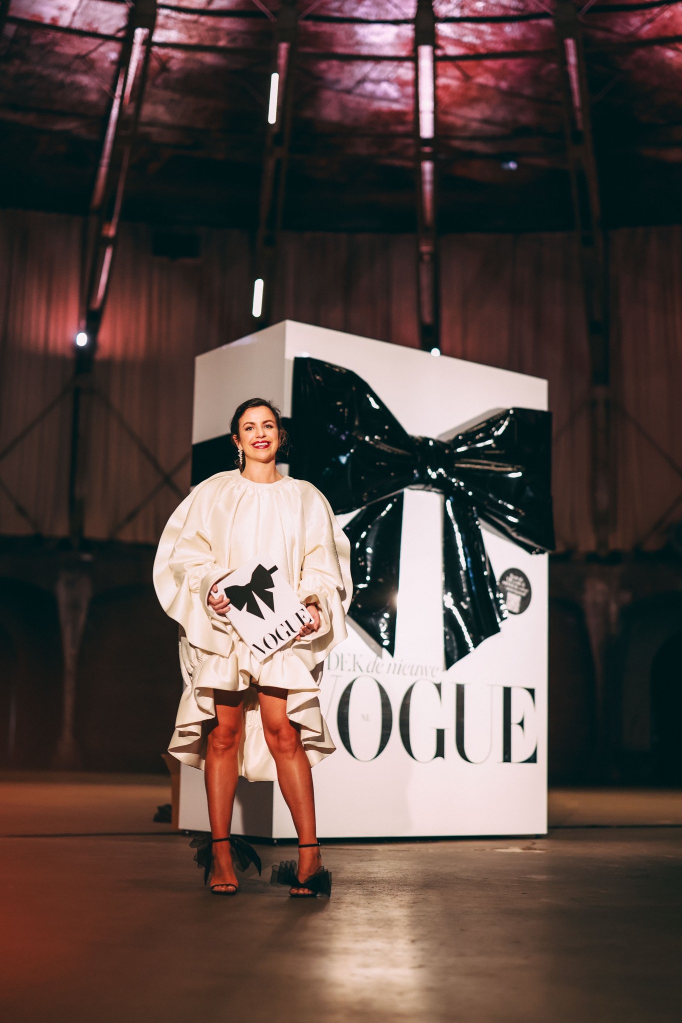 A woman in a white ruffled dress holding the new VOGUE in front of the bow box, in a brick hall