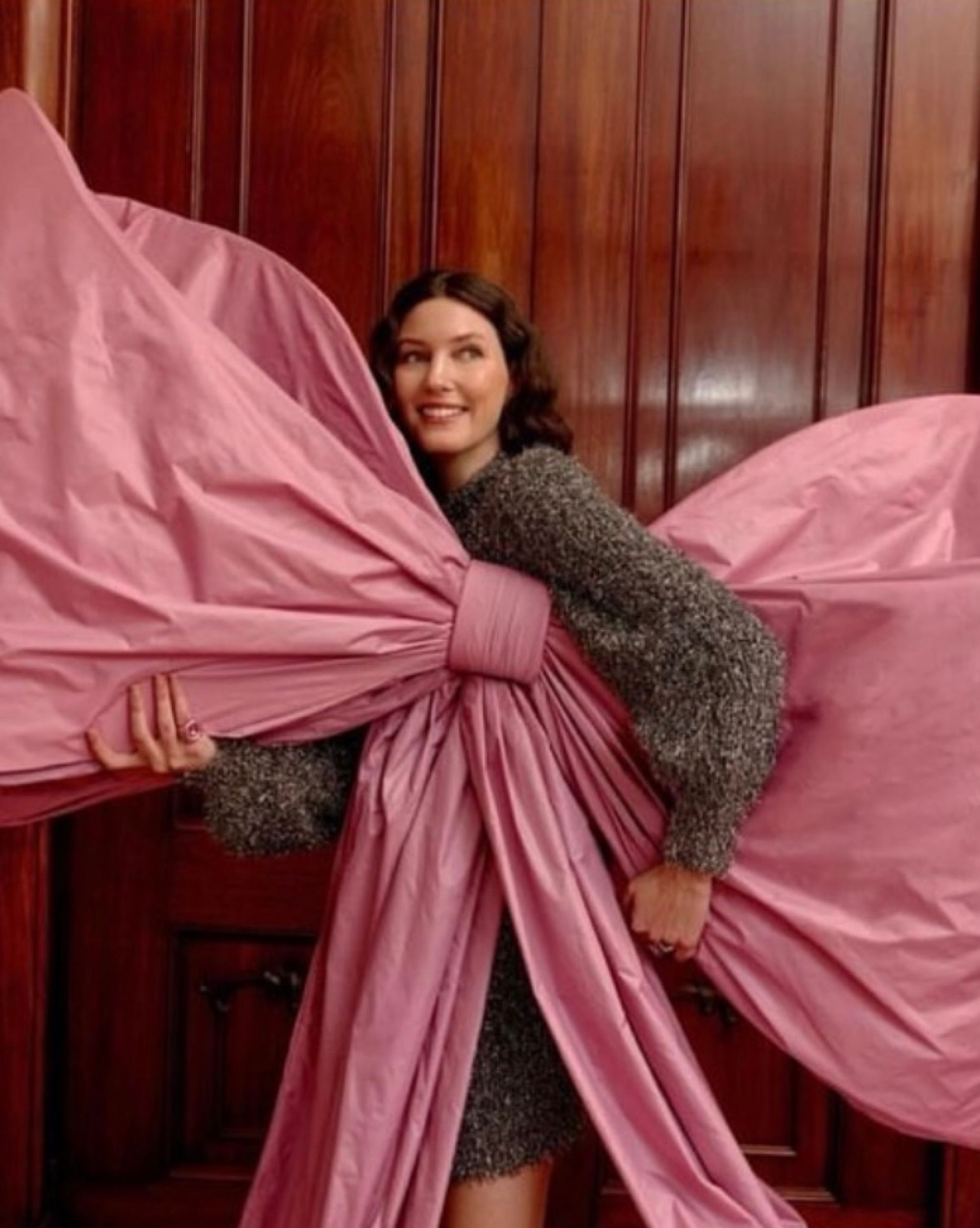 A woman wearing an oversized pink bow, its loops spread wide, in front of wood panelling