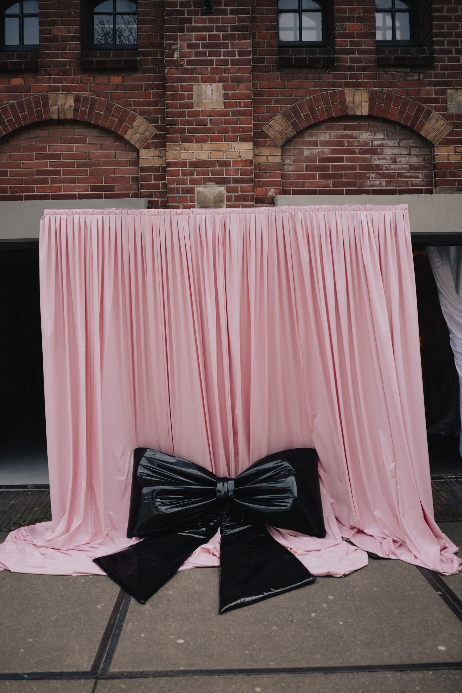 Pink laque drape with a black latex bow against a brick facade