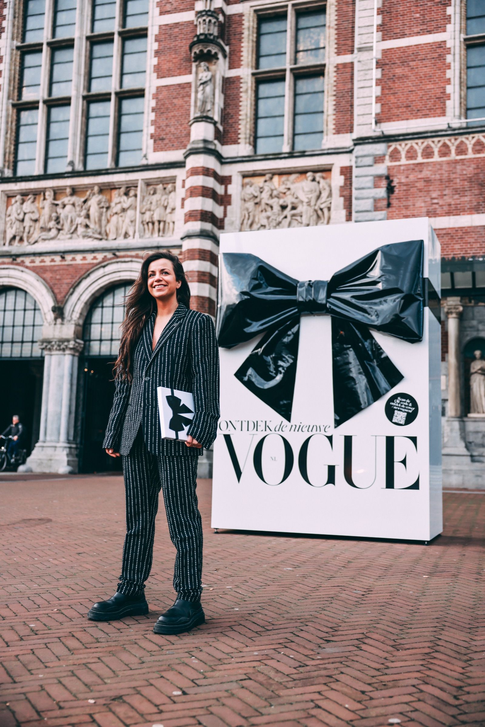 A woman in a pinstripe suit holding the new VOGUE in front of a giant black bow on a white gift box, Rijksmuseum passage