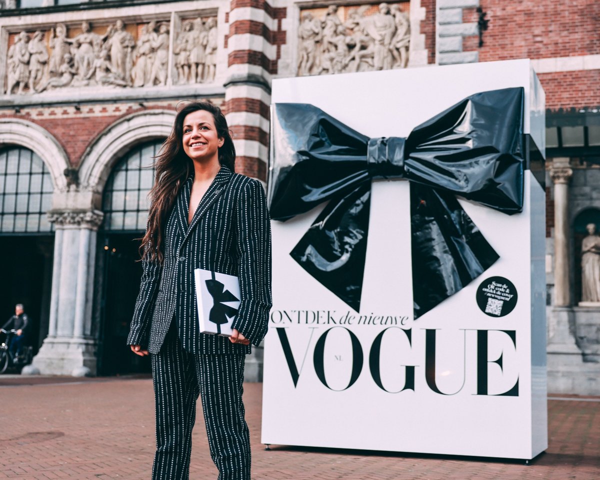 A woman in a pinstripe suit holding the new VOGUE in front of a giant black bow on a white gift box, Rijksmuseum passage