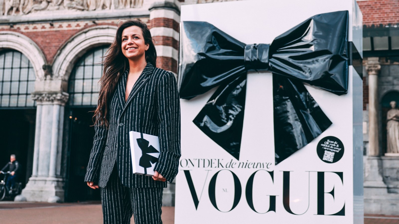 A woman in a pinstripe suit holding the new VOGUE in front of a giant black bow on a white gift box, Rijksmuseum passage