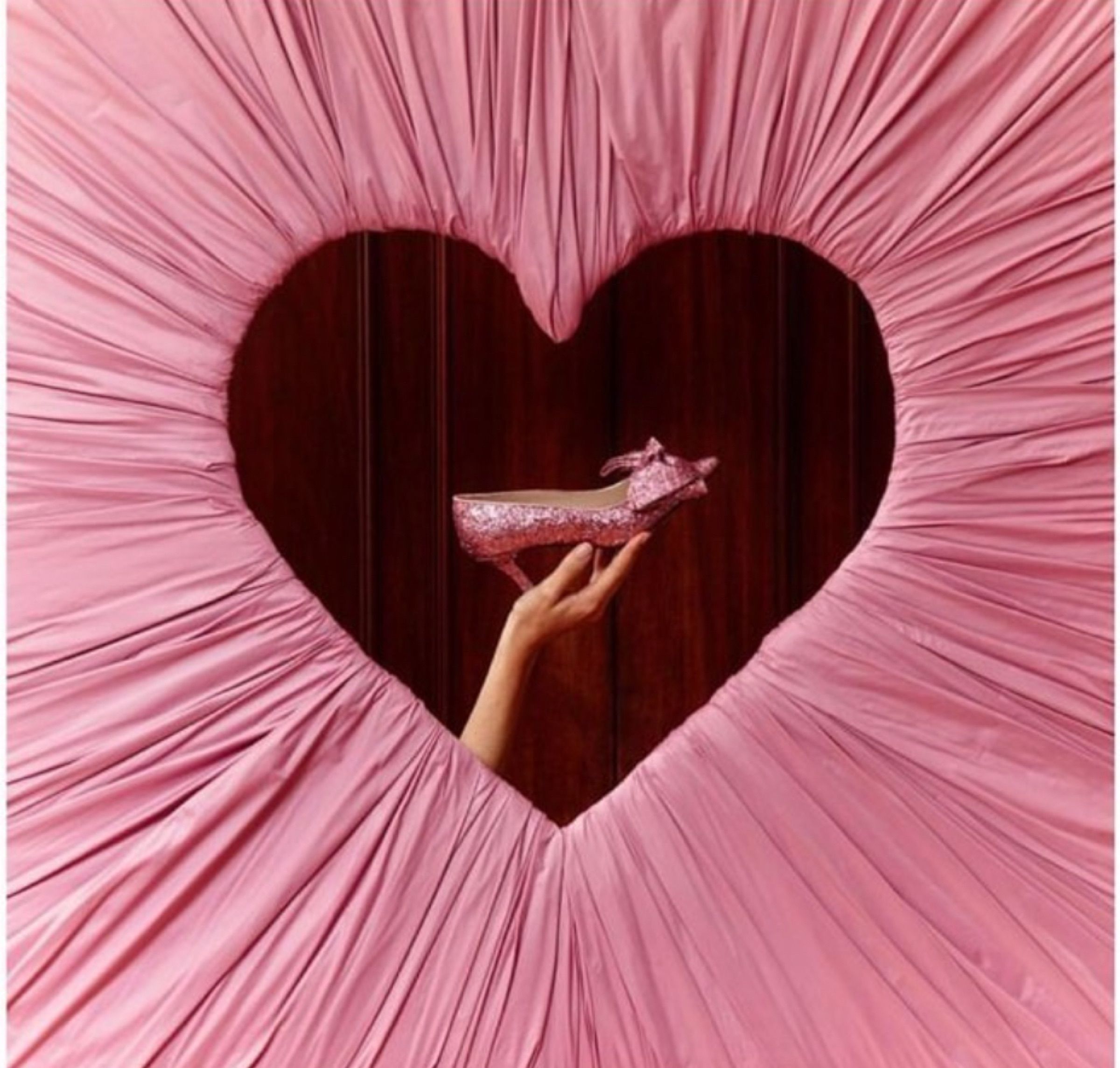 A pink gathered drape cut into a heart, a hand holding a glitter shoe in the opening