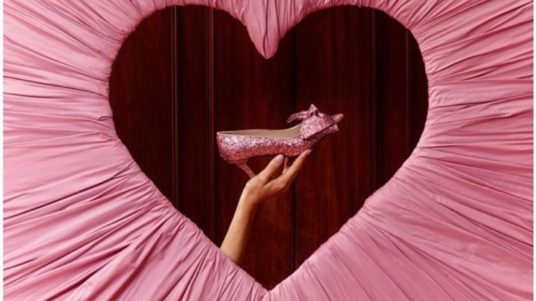 A pink gathered drape cut into a heart, a hand holding a glitter shoe in the opening