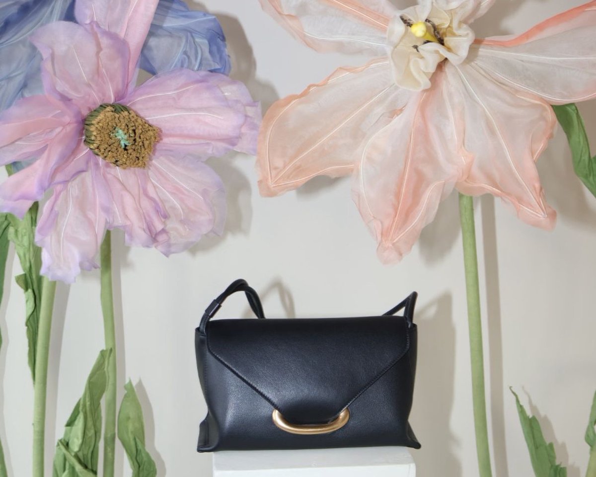 A black Wandler bag on a white plinth between oversized fabric flowers