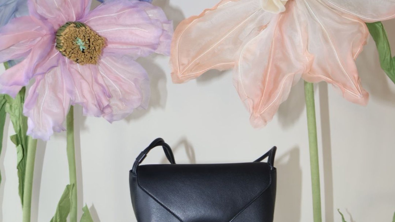 A black Wandler bag on a white plinth between oversized fabric flowers
