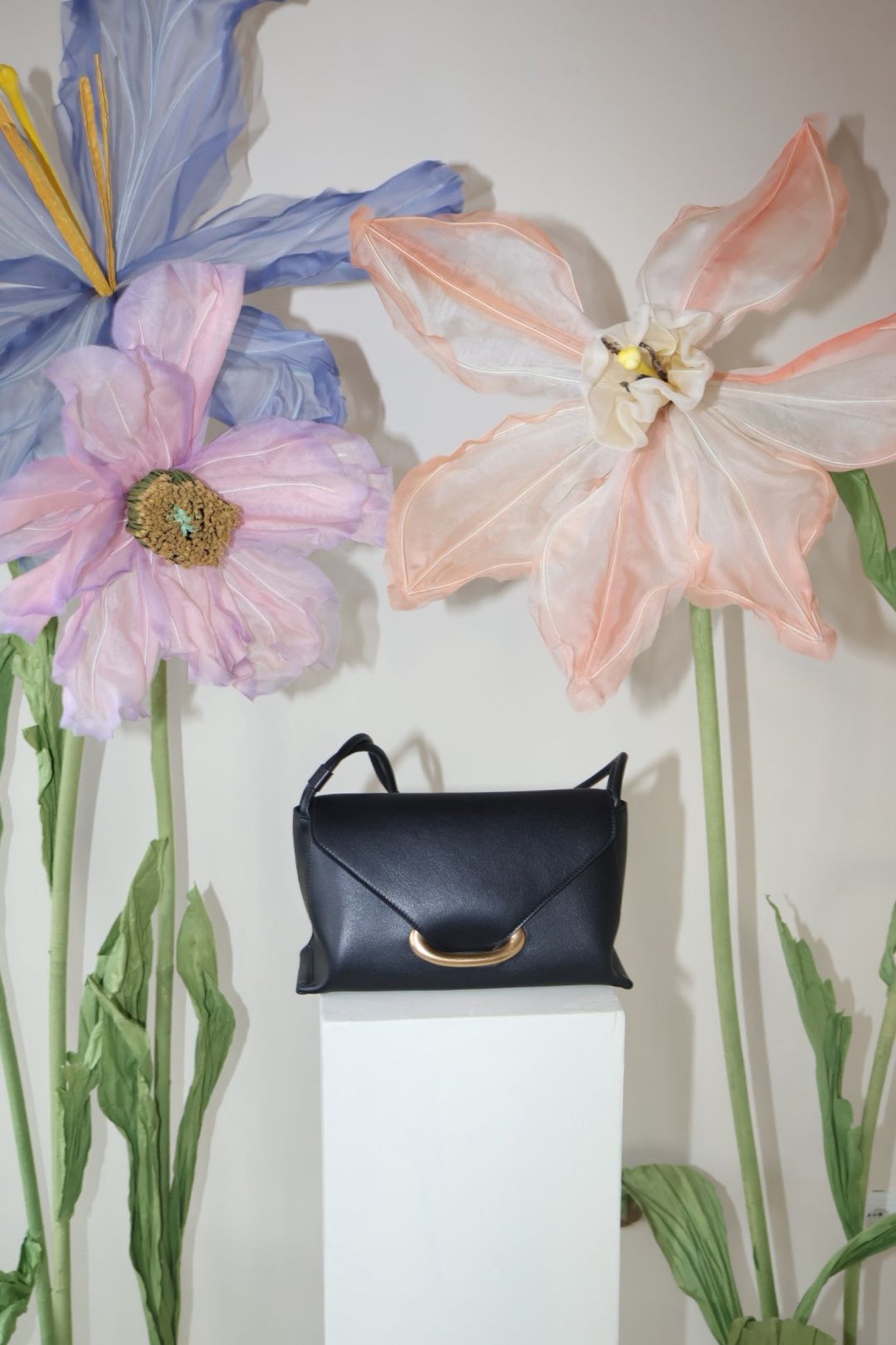 A black Wandler bag on a white plinth between oversized fabric flowers