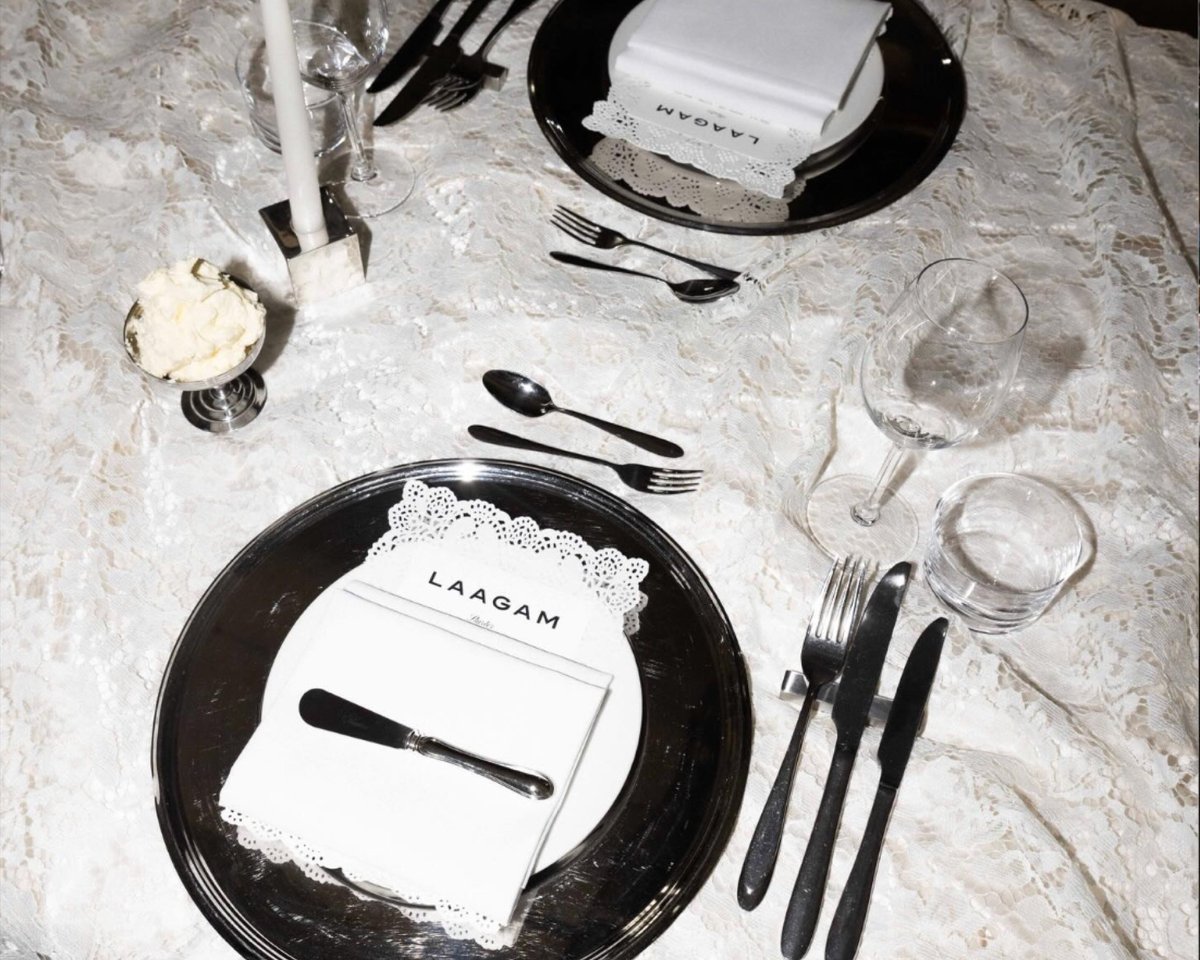 A place setting on white lace: mirror charger, folded napkin with the Laagam card, black cutlery and a candle