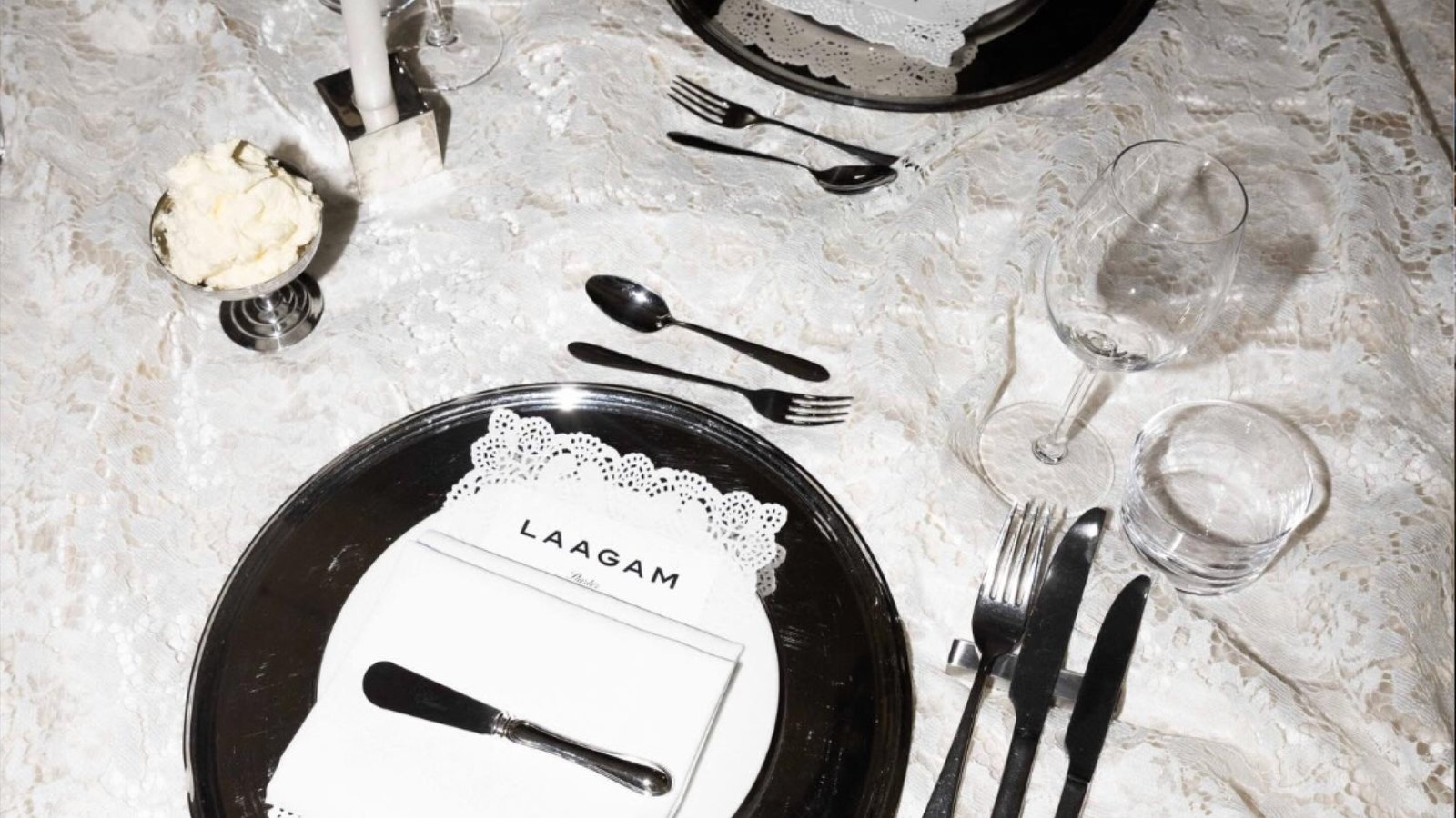A place setting on white lace: mirror charger, folded napkin with the Laagam card, black cutlery and a candle