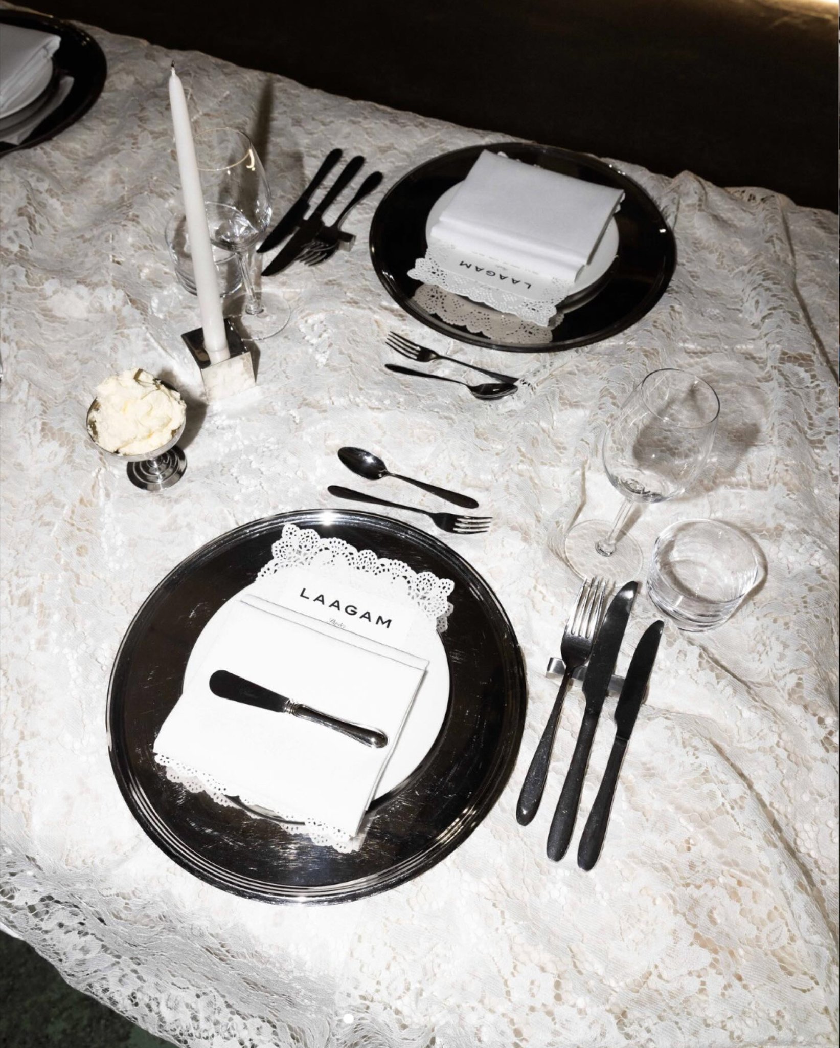 A place setting on white lace: mirror charger, folded napkin with the Laagam card, black cutlery and a candle
