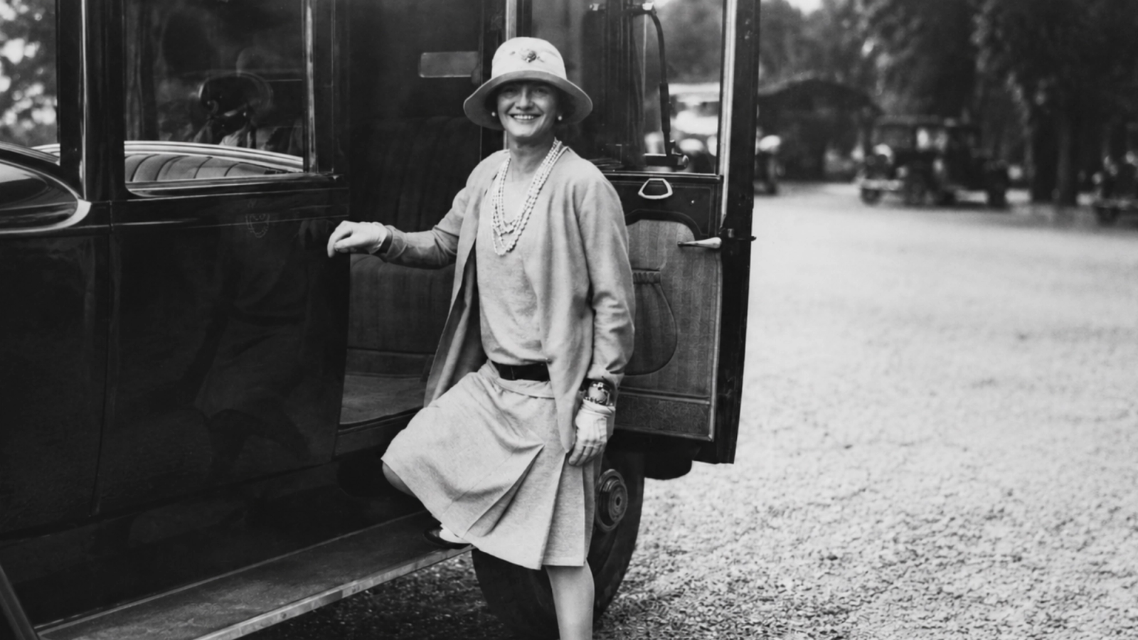 Schwarz-Weiß-Foto einer lächelnden Frau mit Hut und Kleid, die aus einem Oldtimer aussteigt.