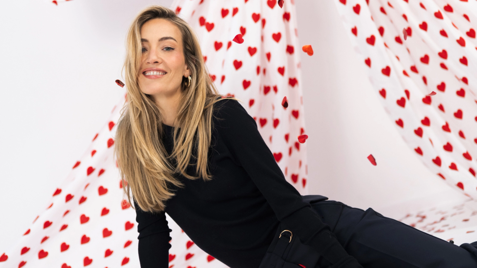 A smiling blonde woman surrounded by red fabric with a heart pattern and falling heart-shaped confetti.
