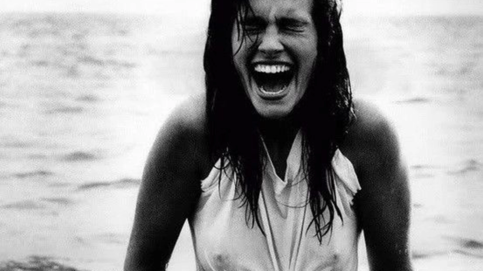Black-and-white photo of a woman with wet hair and a wet shirt, laughing exuberantly with her eyes closed in the water.