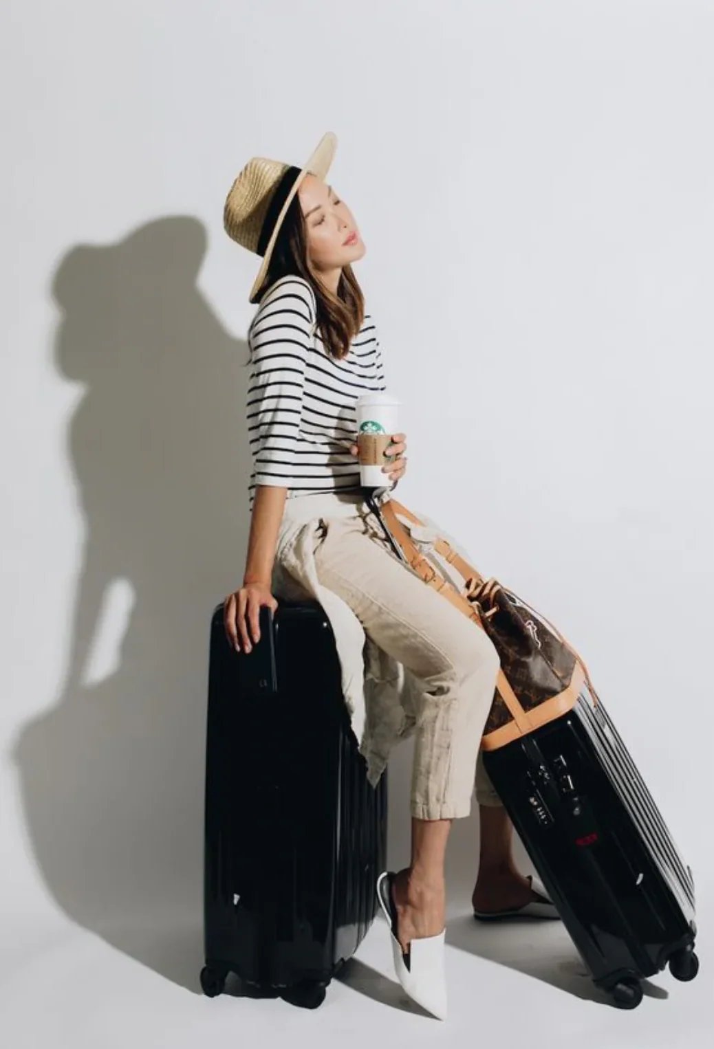 A woman in a straw hat and striped shirt sits on a black suitcase, eyes closed, holding a Starbucks cup, with a Louis Vuitton duffel bag on another suitcase next to her.