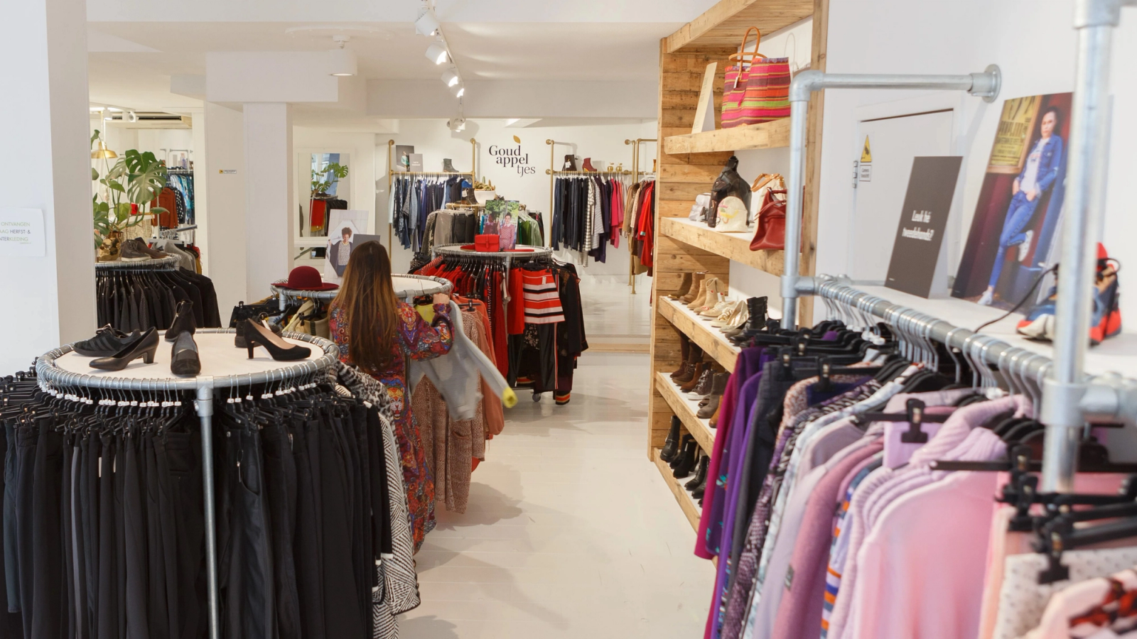 A woman browses racks of clothing in a brightly lit apparel store with wooden shelving.