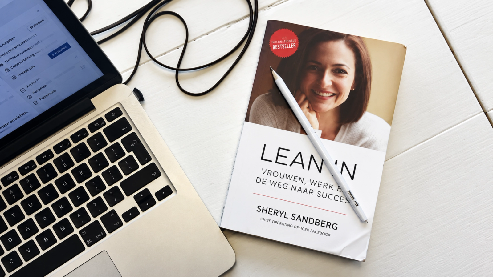 A silver laptop and Sheryl Sandberg's book *Lean In*, with a pen on a white wooden surface.