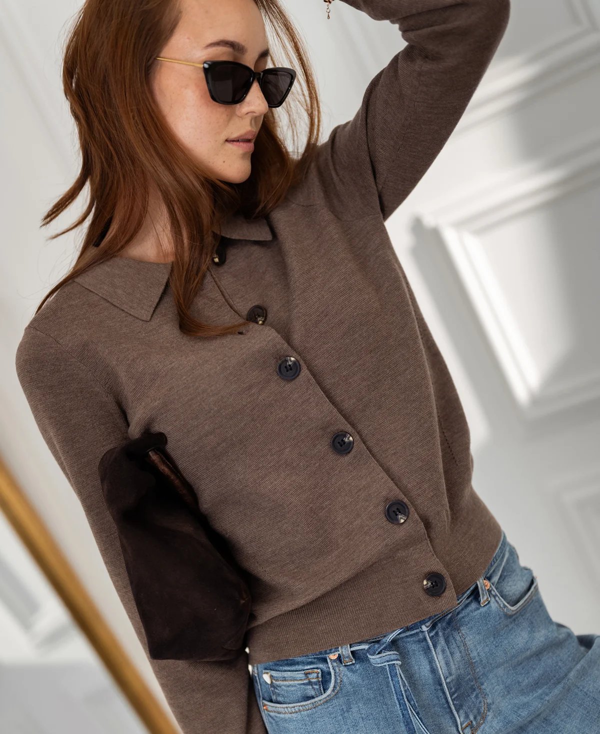 A woman wearing a brown collared cardigan with elbow patches, blue jeans, and black cat-eye sunglasses.