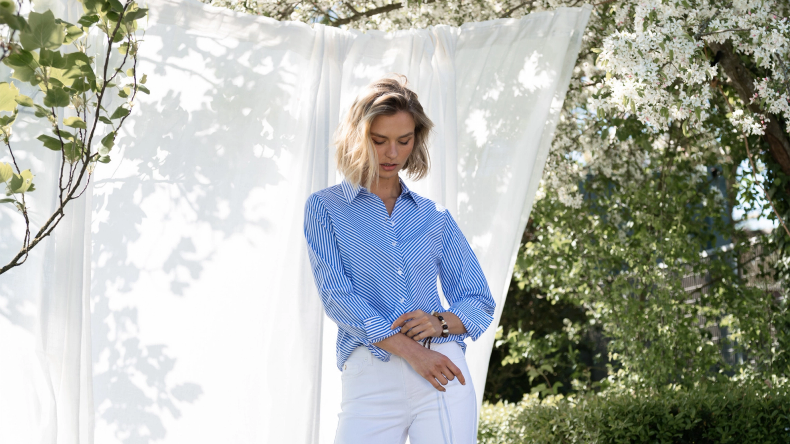 A woman wearing a blue striped shirt and white pants is standing outside against a white backdrop, with flowering trees in the background.