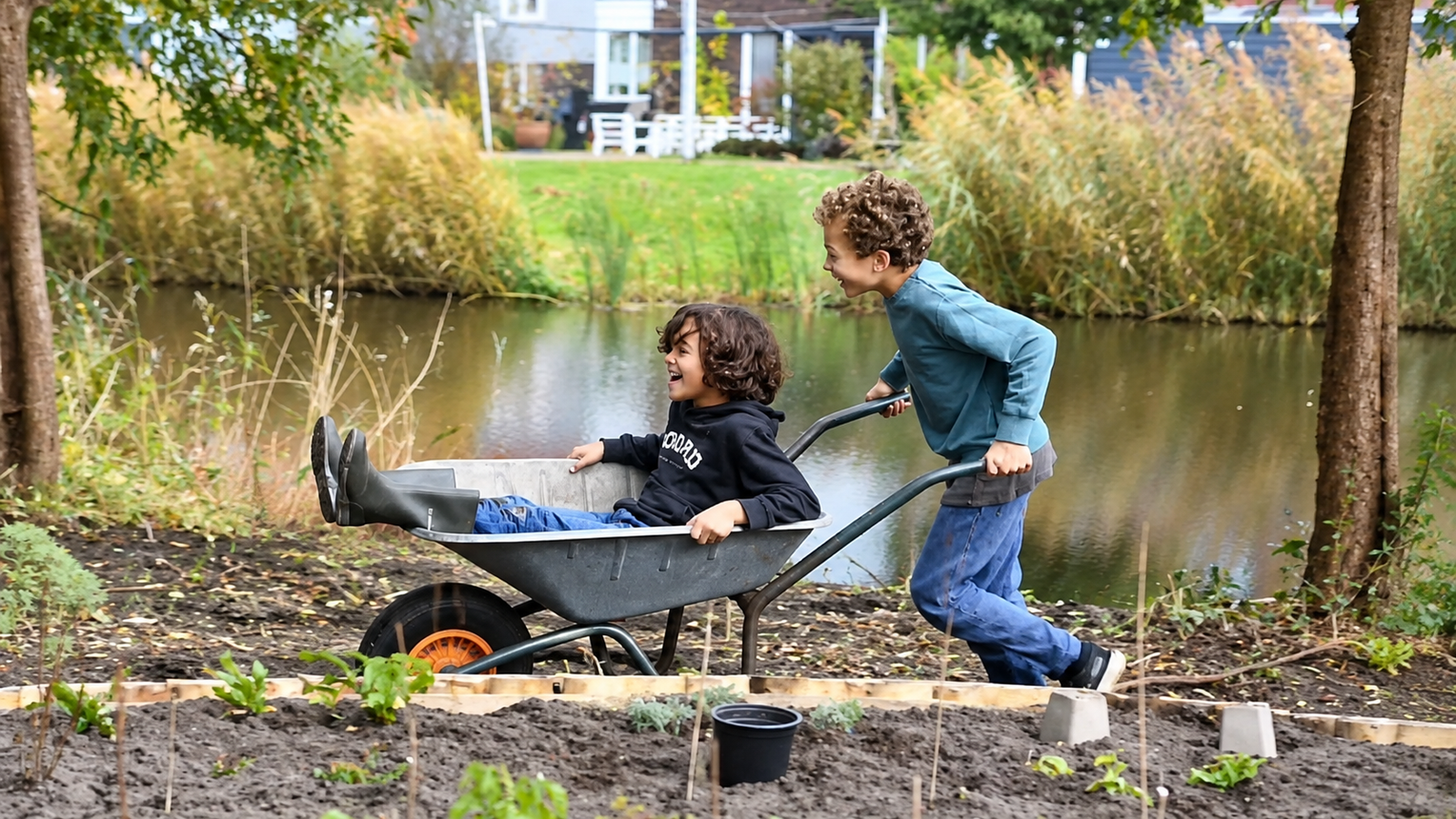 Zwei Jungen lachen, während einer den anderen in einer Schubkarre durch einen Garten am Kanal schiebt.