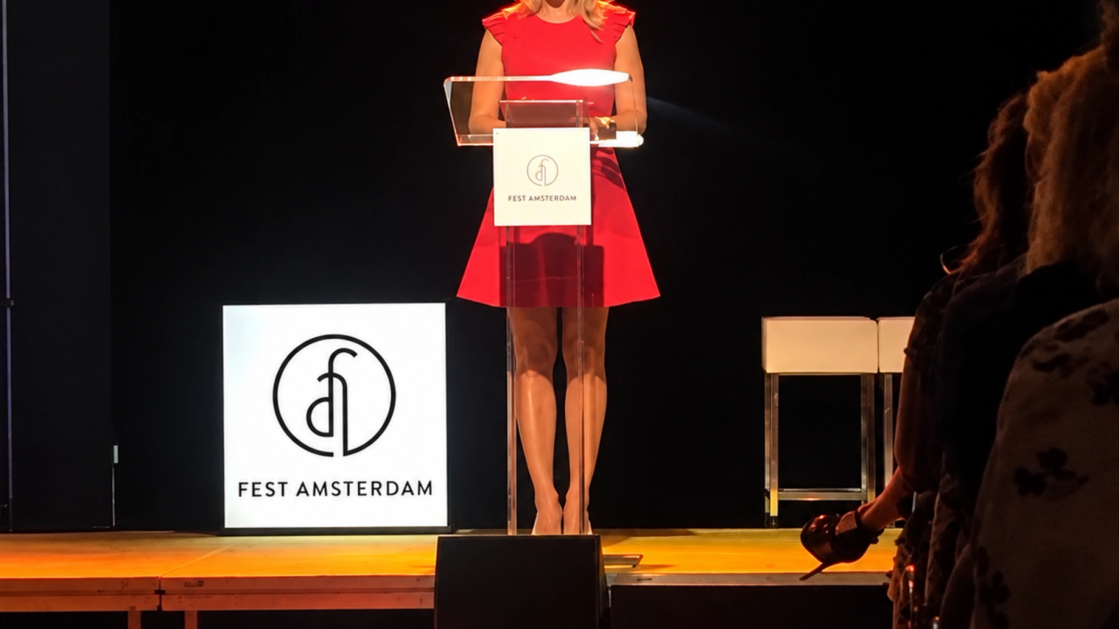 A woman in a red dress is speaking from a podium on a stage, with 