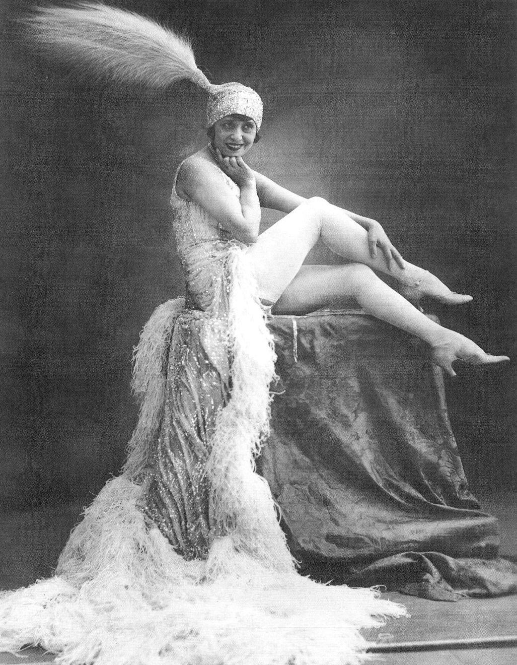 Black-and-white photo of a smiling woman in an elaborate feathered showgirl costume with a plume headdress, seated and posing.