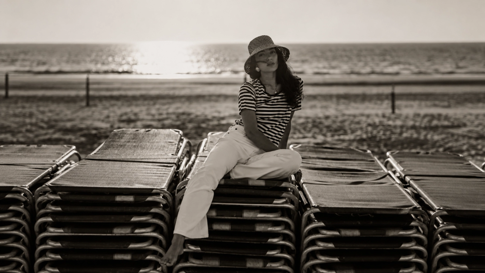 Black and white photo of a woman in a hat sitting on stacked lounge chairs at the beach with the ocean in the background.