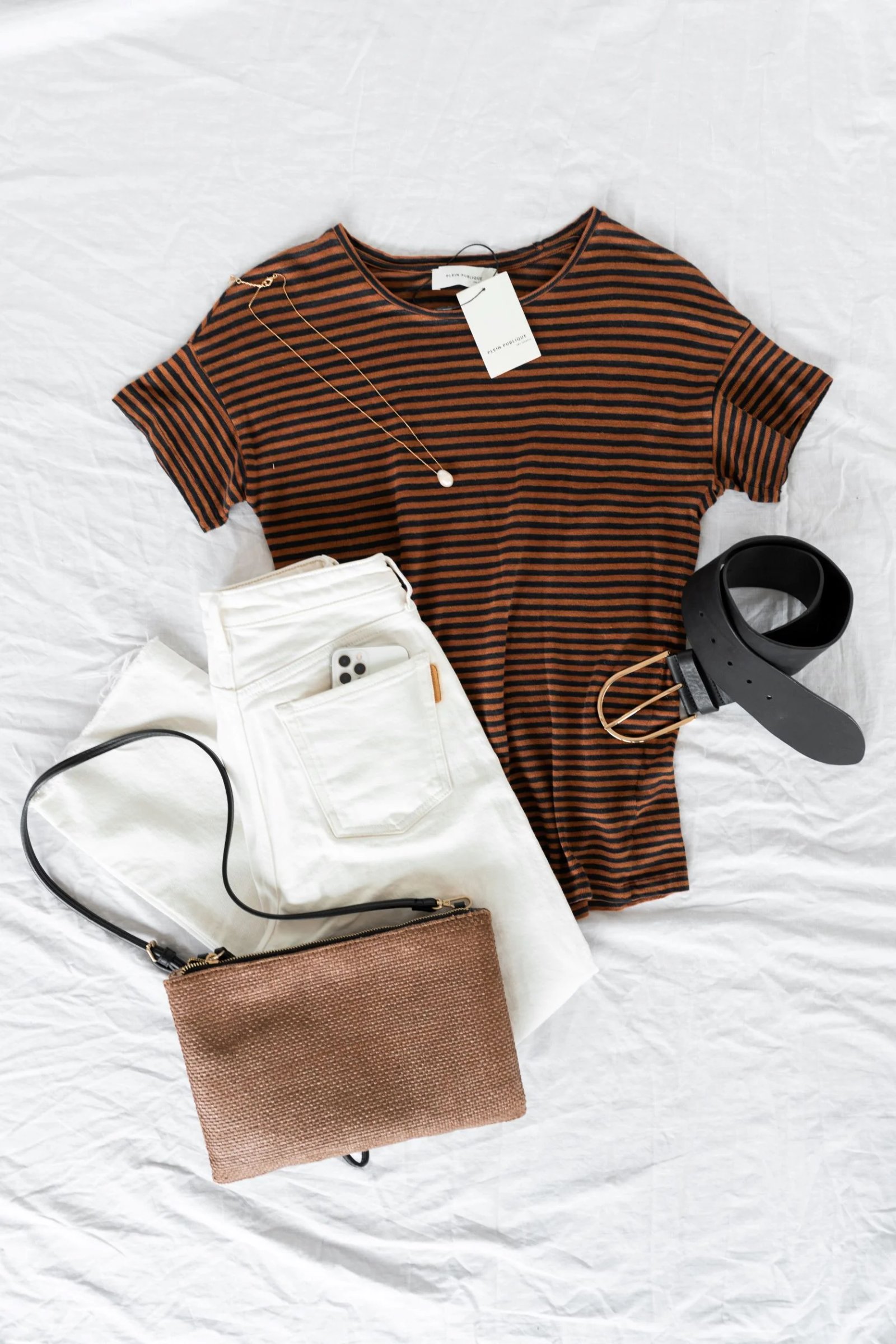 Ein Flatlay zeigt ein braun-schwarz gestreiftes T-Shirt, weiße Jeans, einen schwarzen Gürtel, eine braune gewebte Tasche und eine goldene Perlenkette, die auf einem weißen Hintergrund angeordnet sind.