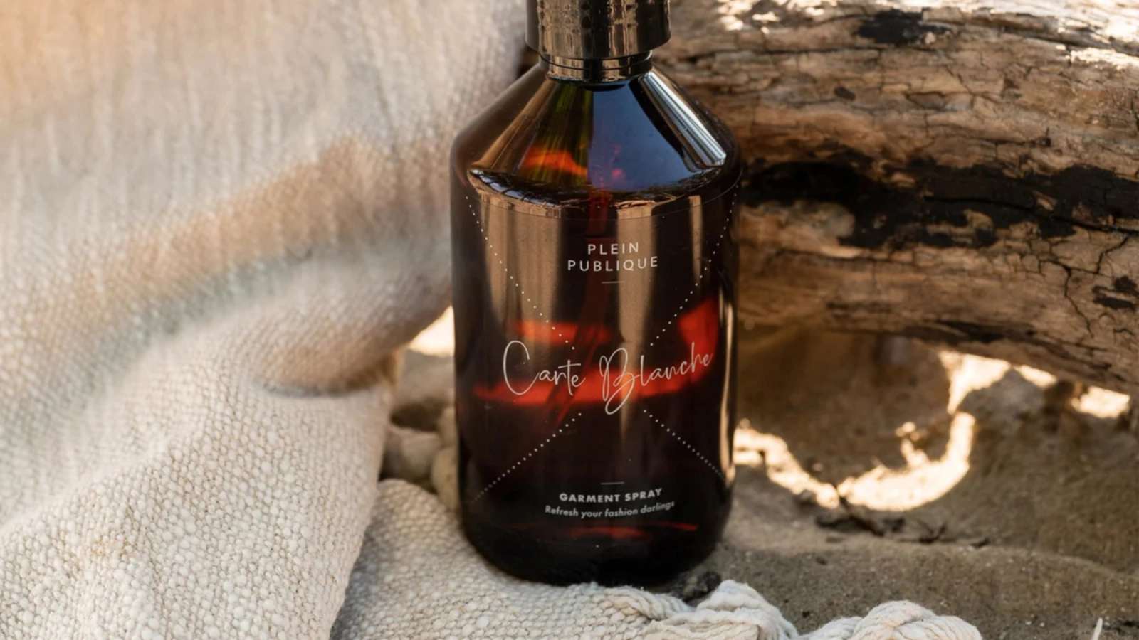 A dark amber bottle of Plein Publique Carte Blanche Garment Spray on light fabric, with sand and driftwood.