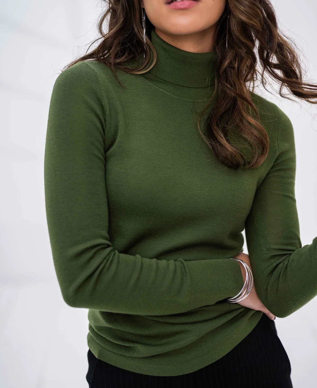 A woman wearing a green turtleneck sweater, black pants, and silver jewelry.