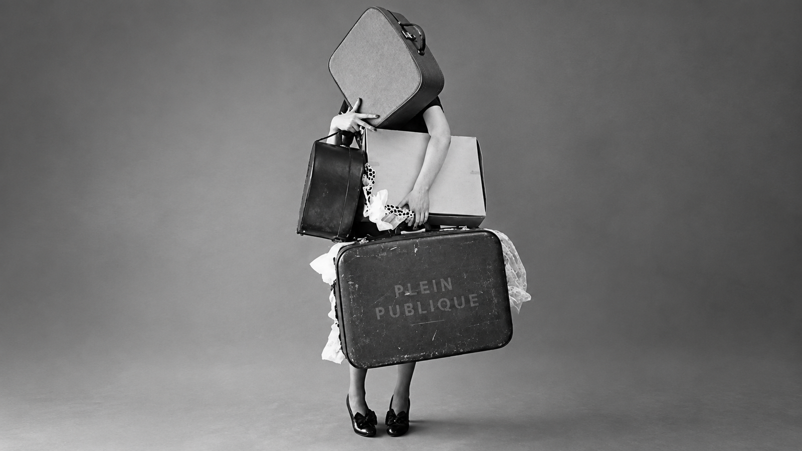 Black and white image of a person mostly hidden behind a tall stack of vintage suitcases.