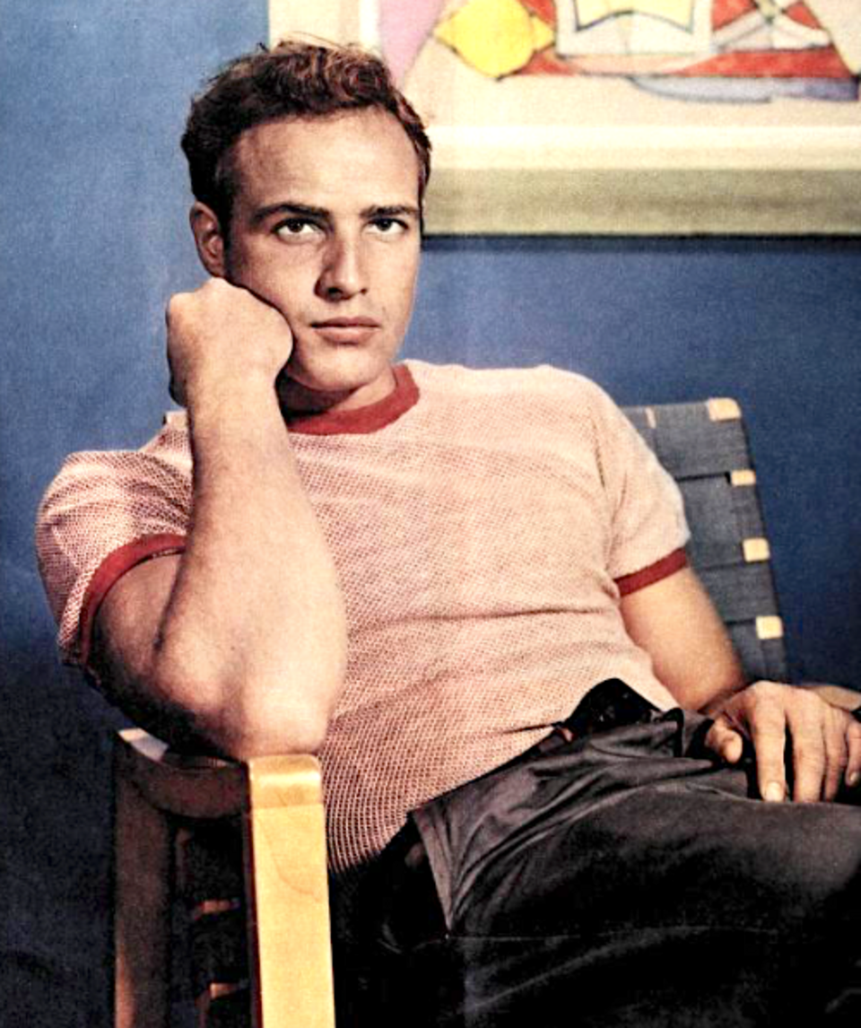 Young Marlon Brando sits in a chair, resting his chin on his hand, wearing a light pink t-shirt.