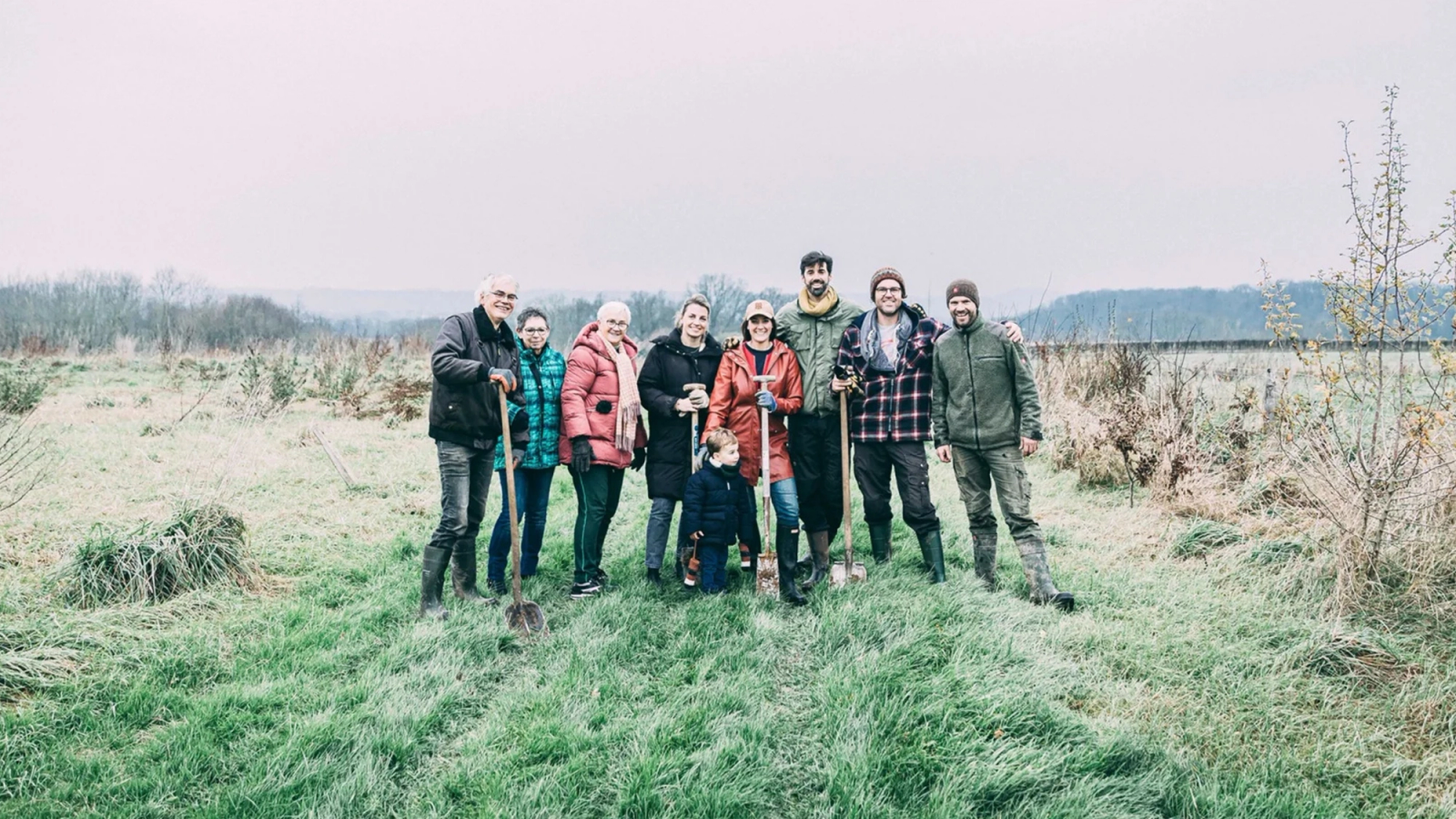 A group of adults and a child stand with shovels in a grassy field with young trees.